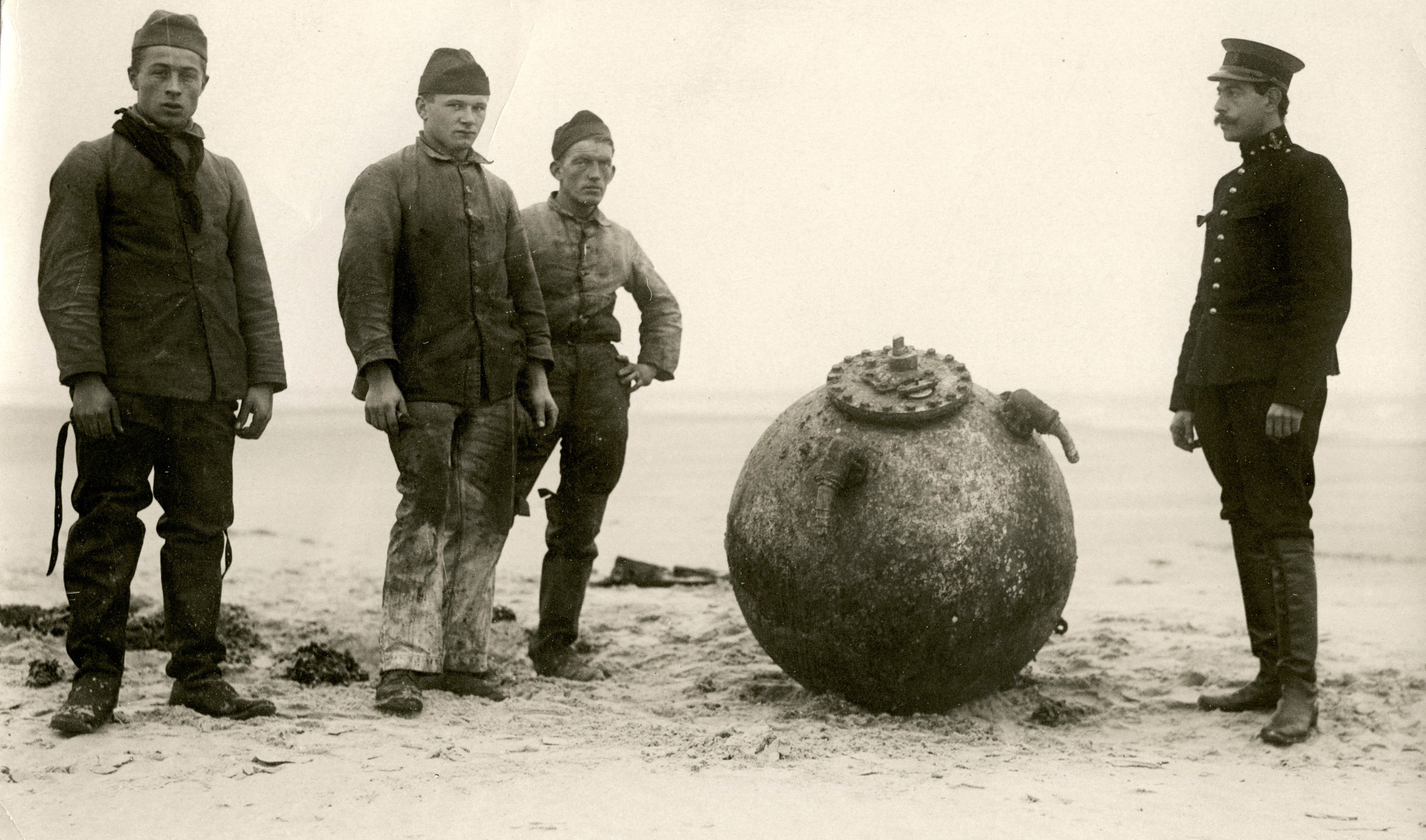 Omschrijving: Een nog niet gedemonteerde landmijn (de voelhoorns zijn nog aanwezig!) aangespoeld op de zandbank Noordland in de mond van de Oosterschelde. De demontage wordt uitgevoerd door het korps Torpedisten onder leiding van 1ste lt. Th. Mollinger (rechts). Foto uit 1918.